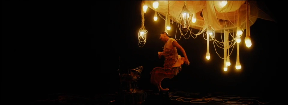 A warm-lit performer moves beneath a cluster of glowing hanging bulbs.
