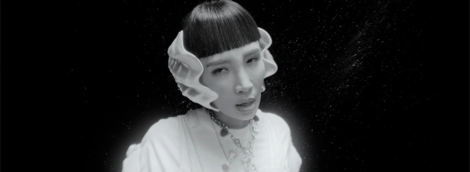 The performer faces the camera in monochrome, with pale ornaments beside her face.