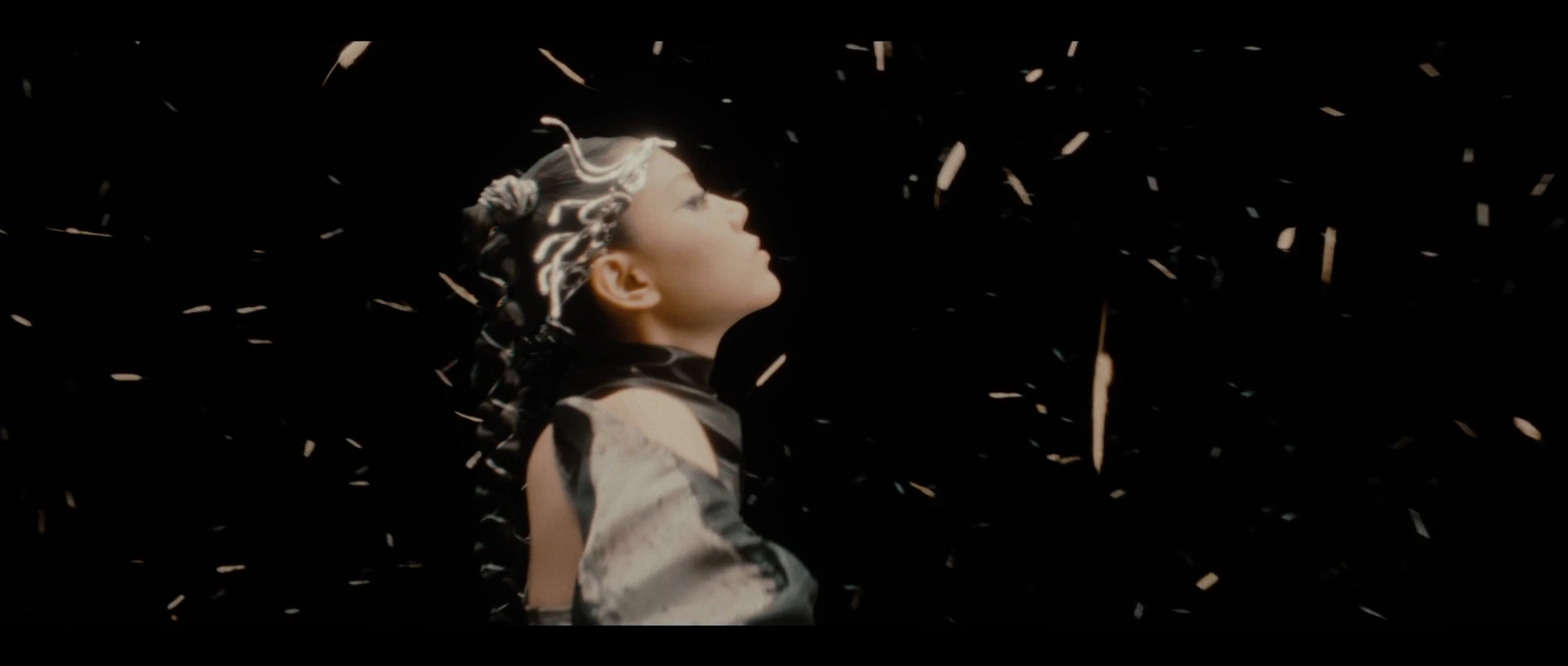 Jasmine Sokko in profile among floating fragments against black.