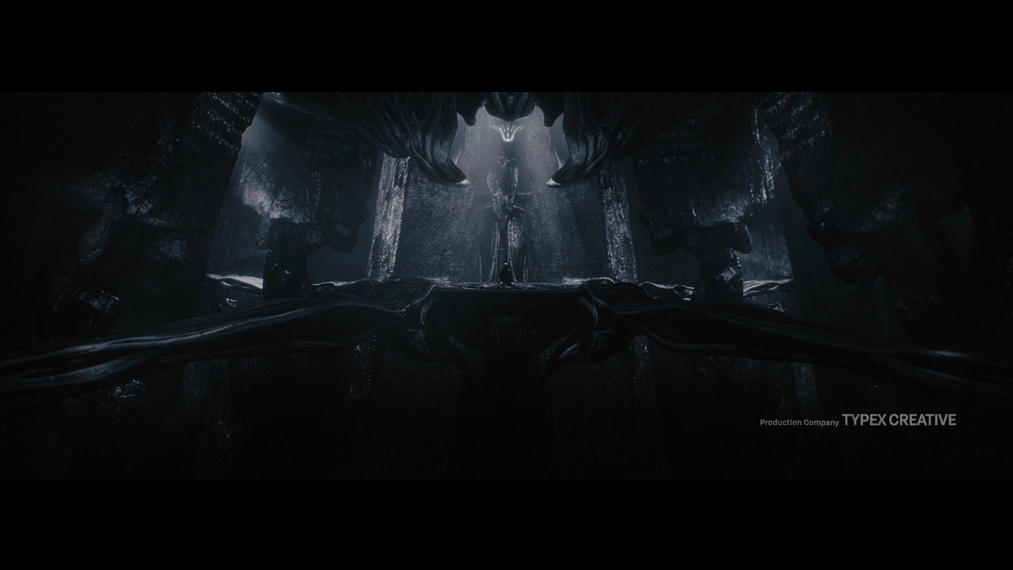 A solitary figure stands within a vast organic chamber, beneath descending sculptural forms.