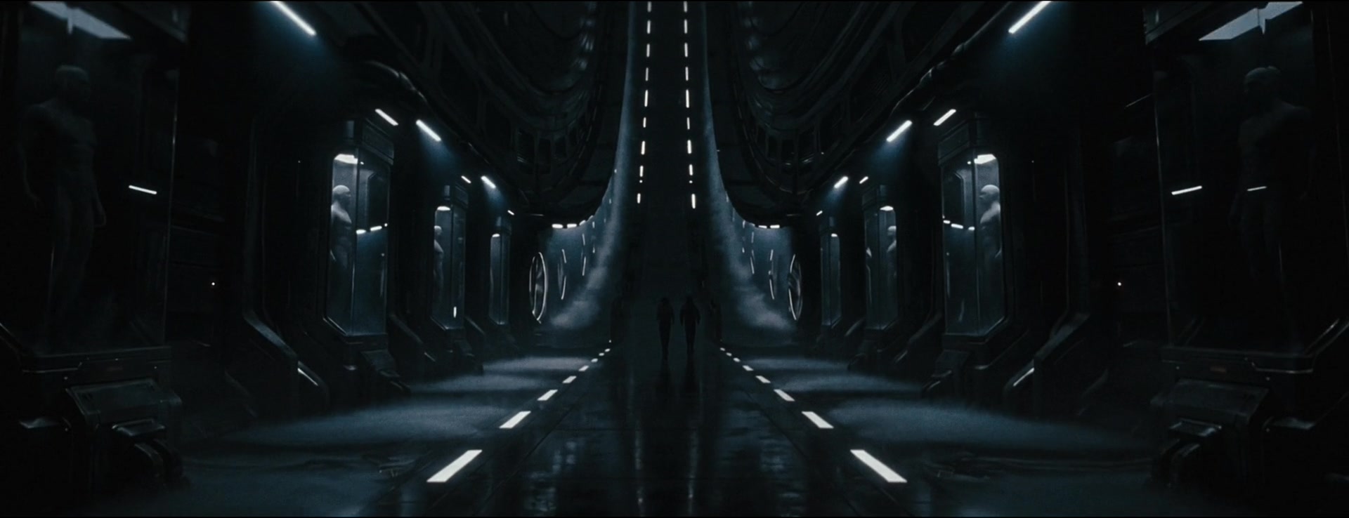 Two figures stand in a soaring laboratory corridor lined with illuminated human chambers.