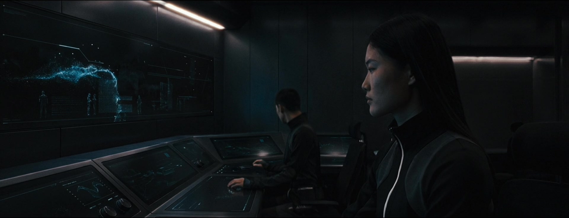A woman in profile monitors a blue holographic display while another operator works behind her.
