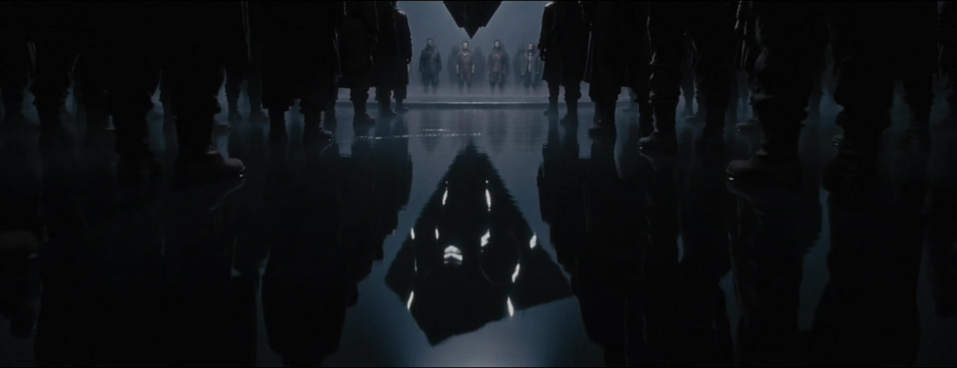A luminous inverted triangle is reflected on wet ground between the feet of a gathered crowd.